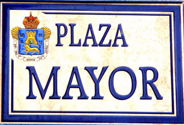 Rechteckige weiße Plakette mit schwarzem Text "Plaza Mayor" in fetter Schrift, blauem Rahmen und einem zentralen gekrönten Wappen-Logo an der Seite eines Gebäudes.