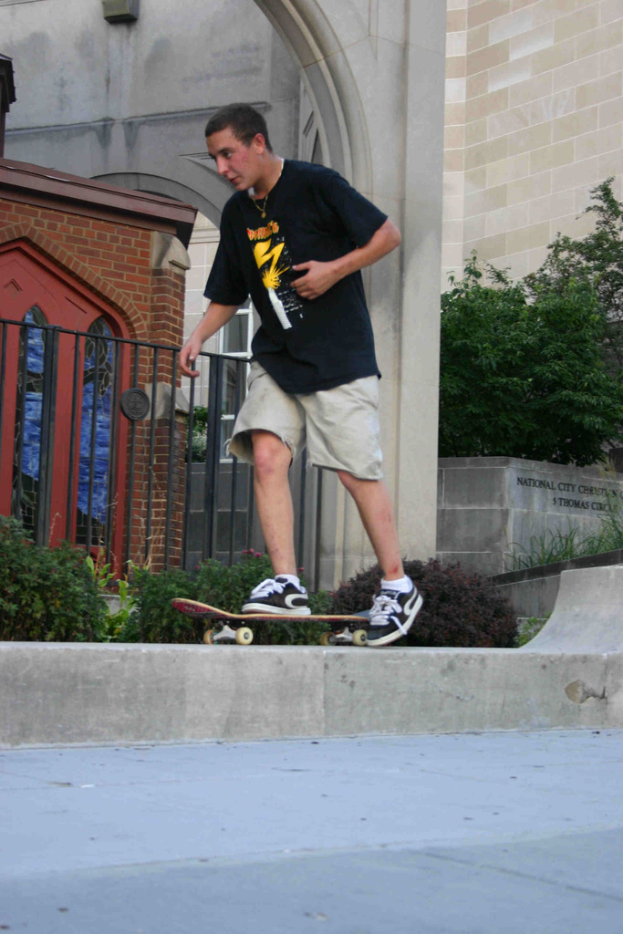 Ein Mann fährt mit einem Skateboard vertikal an einer Wand mit einer Plattform unten, einem Haus, Grill, Pflanzen, Bäumen, einer Backsteinwand und einer Säule im Hintergrund.