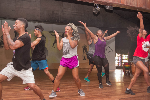 Gruppe von Menschen, die in einem Studio mit einer bemalten Wand, einem Fenster und Deckenleuchten tanzen, wahrscheinlich während eines Zumba-Kurses.