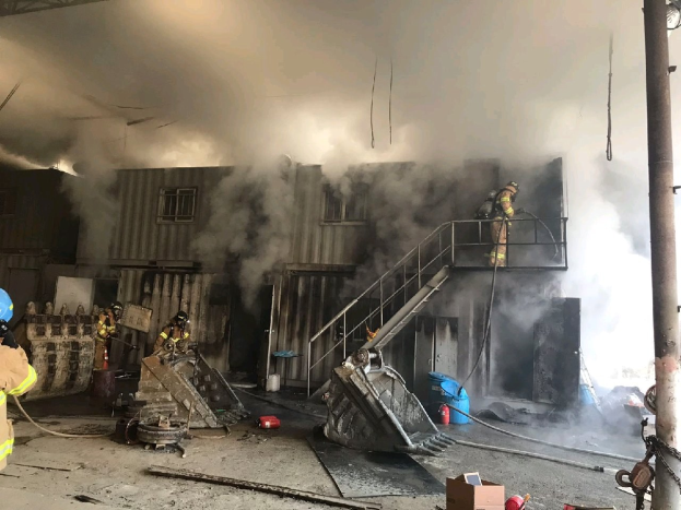 Feuerwehrleute in Helmen verwenden Schläuche, um ein Lagerfeuer zu löschen, mit Rauch, der aus dem Gebäude aufsteigt, Treppenstufen mit Geländern, die zum Eingang führen, eine Kette an einem Pfahl, Fenster im Hintergrund und verstreute Trümmer auf dem Boden.