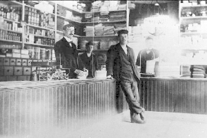 Drei Männer stehen an einem Tresen in einem Laden, umgeben von verschiedenen Gegenständen, mit einem Regal voller Bücher und Gegenstände im Hintergrund, scheinbar mitten in einem Raubüberfall.