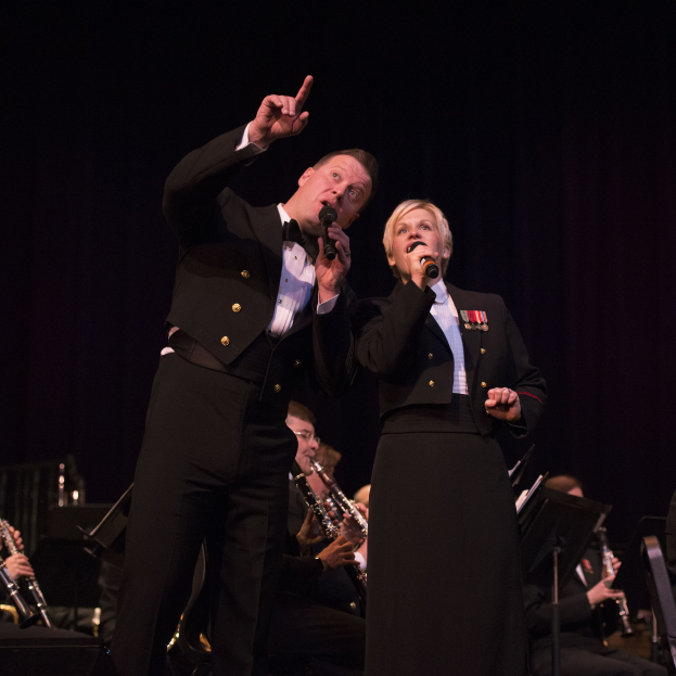 Ein Mann und eine Frau in Smokings stehen auf einer Bühne und halten Mikrofone, während eine Gruppe von Musikern hinter ihnen in einem dunklen Setting sitzt.