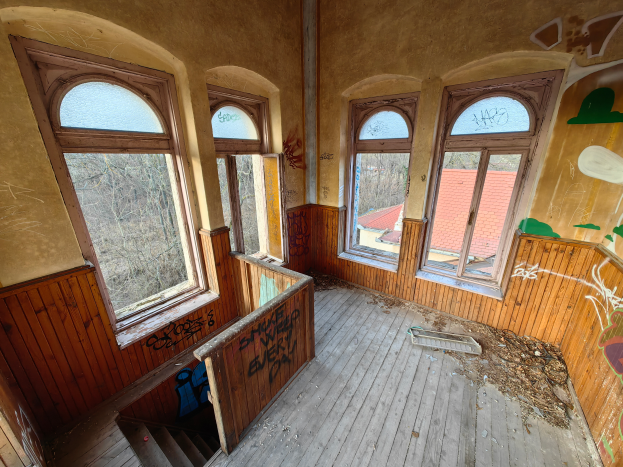 Ein verlassenes Gebäude mit Graffiti an den Wänden und Fenstern, einem Holzfußboden, einer Treppe und verstreuten Gegenständen, mit Bäumen und einem Haus im Hintergrund.