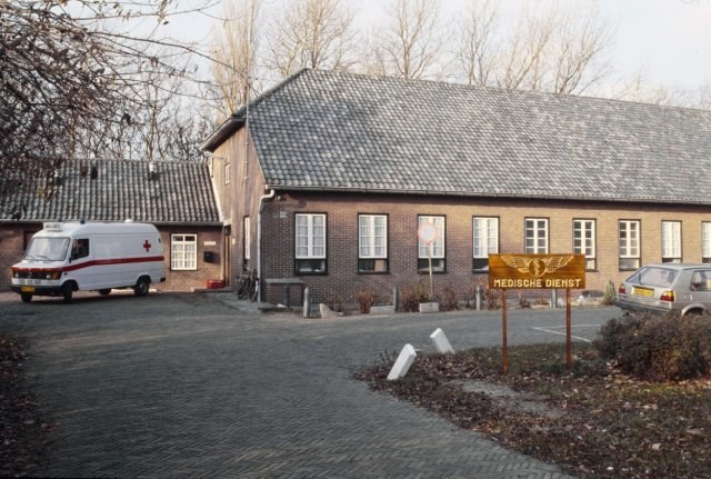Ambulanz vor einem Backsteingebäude mit Fenstern, einer Schildertafel, Pflanzen, Bäumen und einem klaren blauen Himmel geparkt.