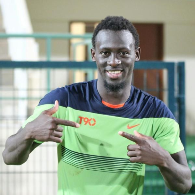 Ein Mann in einem grün-blauen Hemd, der lachend in die Kamera zeigt und im Hintergrund eine Brüstung, eine Wand und eine Tür zu sehen sind.