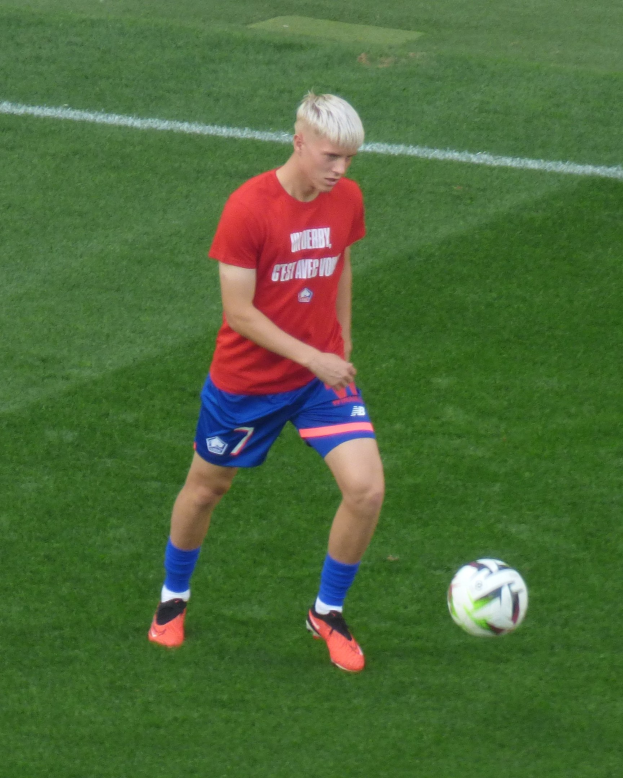 Junger Mann in rotem T-Shirt und blauen Shorts beim Fußballspielen auf einem grünen Rasen, trägt Fußballschuhe und -schuhe während eines Spiels.