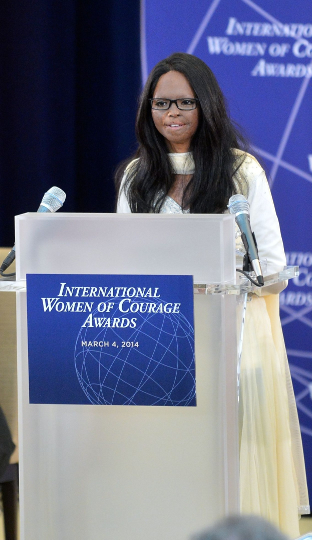 Frau in weißem Kleid und Brille vor Mikrofonen und einer Tafel stehend, bei der 'International Women of Courage Awards 2014'-Veranstaltung mit einem blauen Banner im Hintergrund.