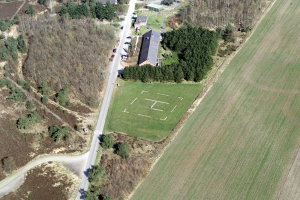 Eine Luftaufnahme eines Bauernhofs inmitten eines Feldes, umgeben von Bäumen, Häusern, Fahrzeugen, Straßen und Gras.