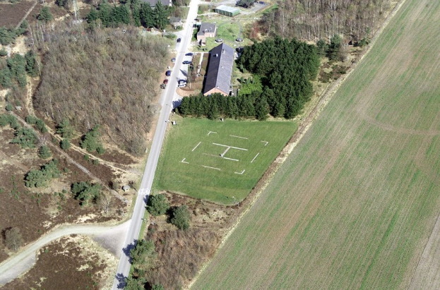 Eine Luftaufnahme eines Bauernhofs inmitten eines Feldes, umgeben von Bäumen, Häusern, Fahrzeugen, Straßen und Gras.