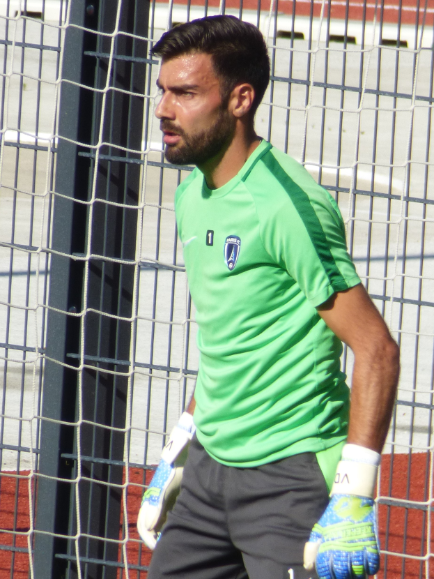 Ein Mann in einem grünen Hemd und grauen Shorts steht neben einem Fussballtor und trägt einen Handschuh, was darauf hinweist, dass er ein professioneller Fussballspieler ist.
