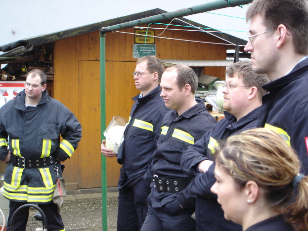 Eine Gruppe von Männern in blauen Feuerrettungsuniformen, die nach links schauen, mit einem kleinen hölzernen Schuppen und Rohrrahmen im Hintergrund.