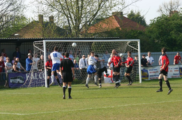 Spieler spielen Fußball auf einem Feld mit einem Tor hinter ihnen, während Zuschauer von der Seitenlinie zusehen, mit Bäumen und Häusern im Hintergrund.