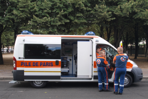 Ambulanz am Straßenrand mit drei Personen davor geparkt, umgeben von Bäumen, Gebäuden und anderen Fahrzeugen.