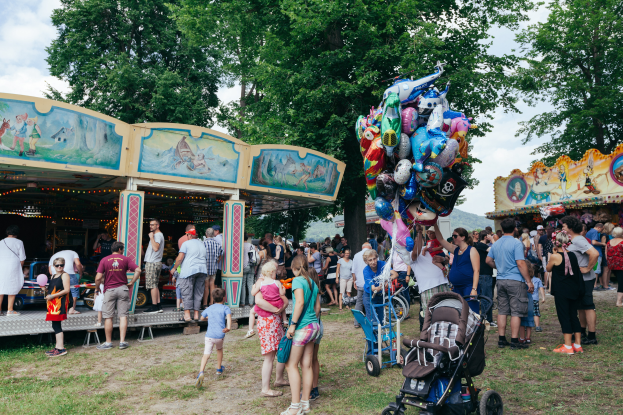 Eine Menschenmenge, einige mit Kinderwagen, versammelte sich um ein bunt beleuchtetes Karussell auf einem Volksfest, mit Bäumen und einem bewölkten Himmel im Hintergrund.