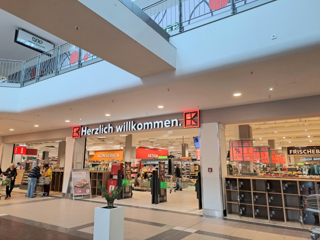 Das Innere eines Einkaufszentrums mit Menschen, die herumlaufen, Geschäften, Schildern mit der Aufschrift "Herzlich willkommen", Lampen, Geländern und einer Topfpflanze unten.
