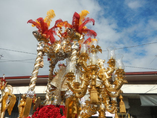 Ein großes goldenes und rotes Festwagen, geschmückt mit Blumen und anderen Dekorationsgegenständen bei einem Karnevalsumzug, mit einem Gebäude, Strommasten mit Drähten und einem bewölkten Himmel im Hintergrund.