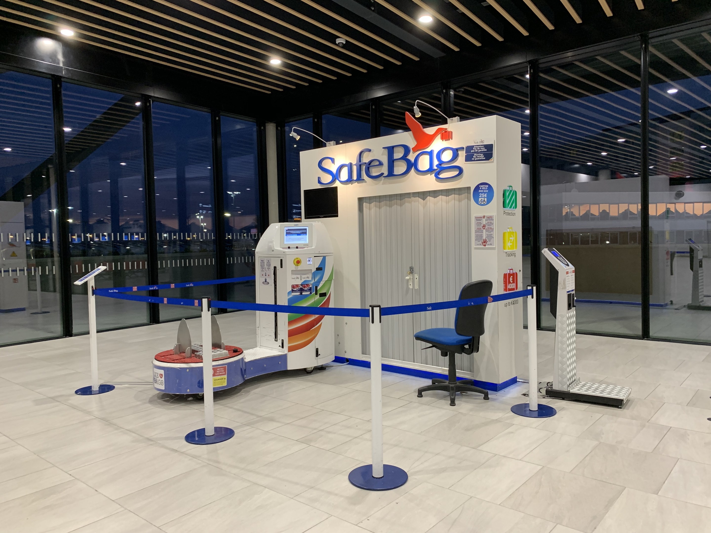 Sicherheitskontrolle am Flughafen mit einem Stuhl, Absperrgittern, Maschinen, einem Bildschirm, einer bedruckten Tafel, Glaswänden, Deckenleuchten und sichtbarem Bodenbelag.