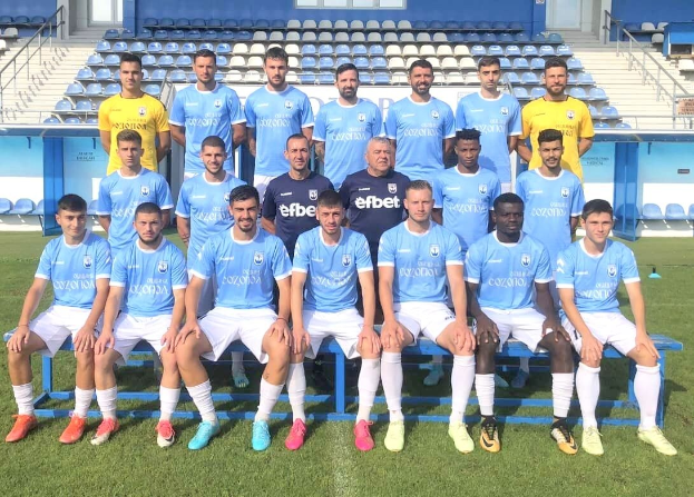 Eine Gruppe von Männern, einige sitzen auf Bänken und andere stehen auf einem Fußballfeld, mit Stühlen, Treppen, Geländern, Wänden und Fenstern im Hintergrund.