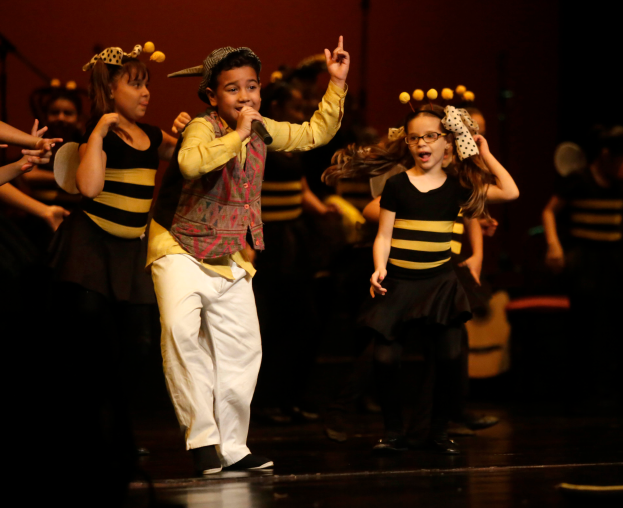 Eine Gruppe von Kindern in Bienenkostümen tanzt auf einer Bühne, ein Junge in der Mitte hält ein Mikrofon, dahinter andere Kinder und eine Wand.