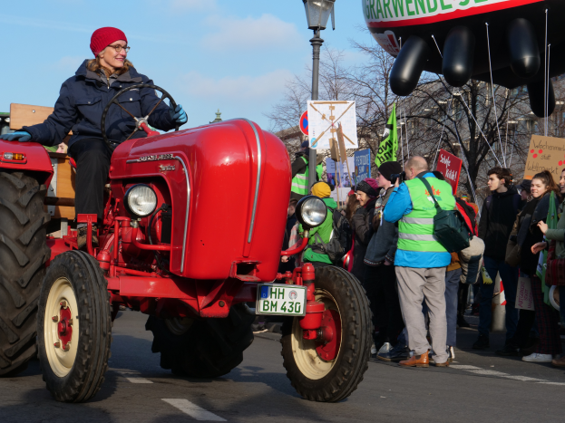 Eine Frau fährt einen roten Traktor auf einer Straße, umgeben von einer Menge mit Schildern und Transparenten, mit Laternenpfählen, Bäumen, Gebäuden und einem klaren blauen Himmel im Hintergrund.