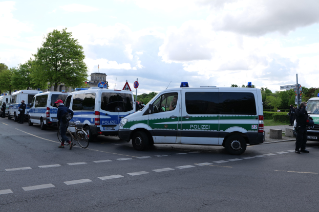 Eine Gruppe von Polizeiwagen, die entlang einer Straße geparkt sind, mit einem Radfahrer im Vordergrund, Bäume und Gebäude im Hintergrund unter einem bewölkten Himmel.