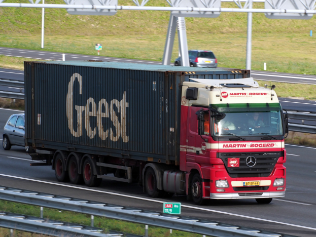 Ein roter Lastwagen mit einem Container auf dem Rücken fährt auf einer Autobahn, mit ein paar Menschen darin, und mit Strommasten, Holzplanken und Gras im Hintergrund.