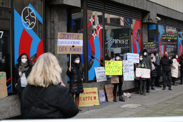 Eine Gruppe von Menschen mit Masken und Schildern protestiert vor einem Gebäude mit Glaswänden und -türen, mit Texttafeln, Kameras und einem Hund.