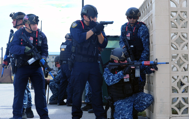 Gruppe von Polizisten in Uniform auf einer Brücke stehend, mit Helmen und Waffen ausgerüstet, mit einer Wand auf der rechten Seite und Gebäuden, Bäumen und Himmel im Hintergrund.