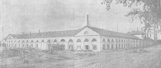 Schwarzes und weißes Foto eines großen, mehrfenstrigen Gebäudes, das von Grün umgeben ist und unter einem klaren Himmel steht, mit der Aufschrift "1892 – das erste Fabrikgebäude der Stadt" unten.