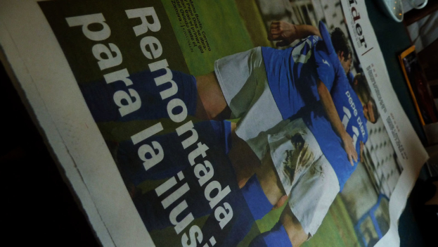 Eine Zeitung auf einem Tisch zeigt drei Männer in blauen T-Shirts, möglicherweise beim Fußballspielen, mit Text am unteren Rand des Bildes.