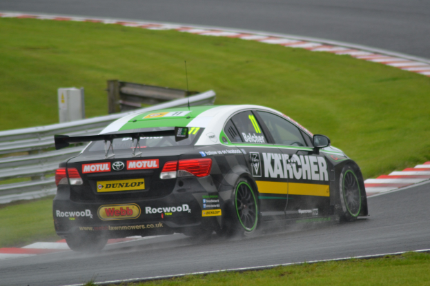 Ein Auto mit Text an der Seite, das auf einer regennassen Rennstrecke fährt, umgeben von Gras, mit einem Geländer und einem hölzernen Objekt im Hintergrund.