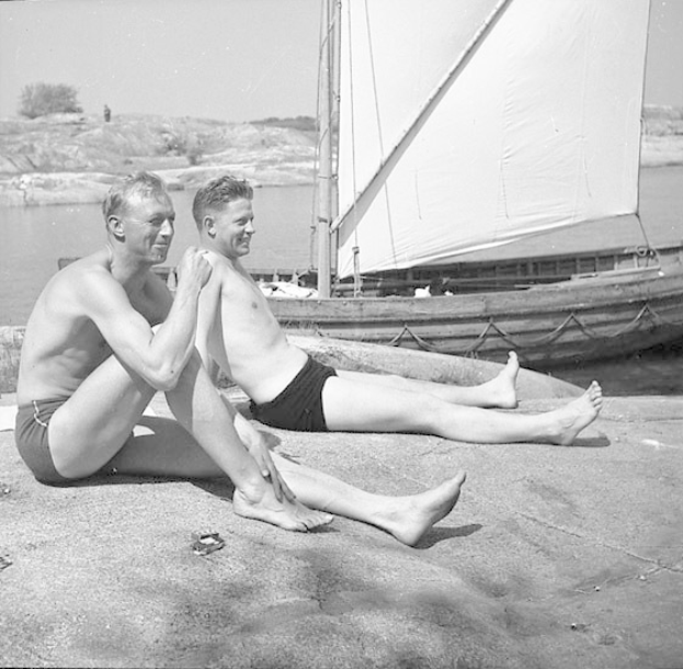 Zwei Männer in Badebekleidung sitzen am Strand neben einem Segelboot, mit blauem Wasser, grünen Bäumen und einem weißen Himmel im Hintergrund.