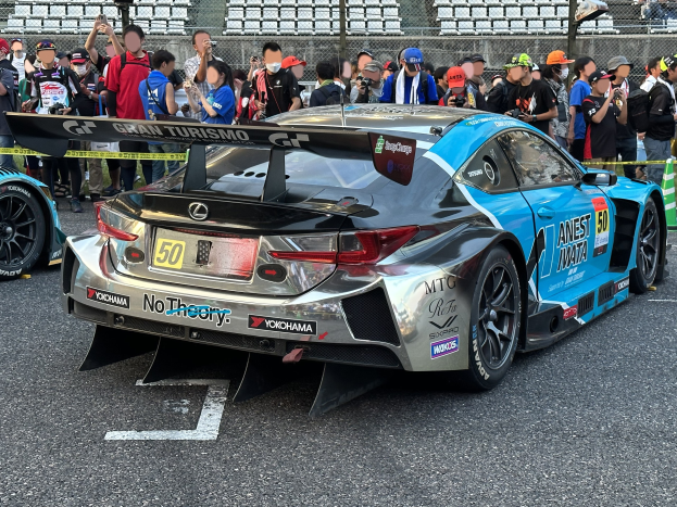 Ein schwarzer Lexus RC F GT3 Rennauto umgeben von Zuschauern mit Kameras und Handys, mit Maschendrahtzaun und Stühlen im Hintergrund.