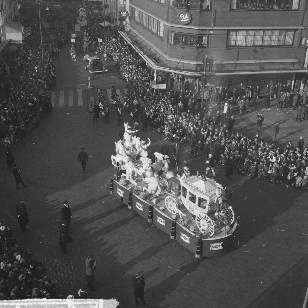 Eine große Menschenmenge umringt einen Festwagen bei einem Umzug, der mit bunten Luftballons und Girlanden geschmückt ist, mit Gebäuden und Laternen im Hintergrund, in Schwarz-Weiß.