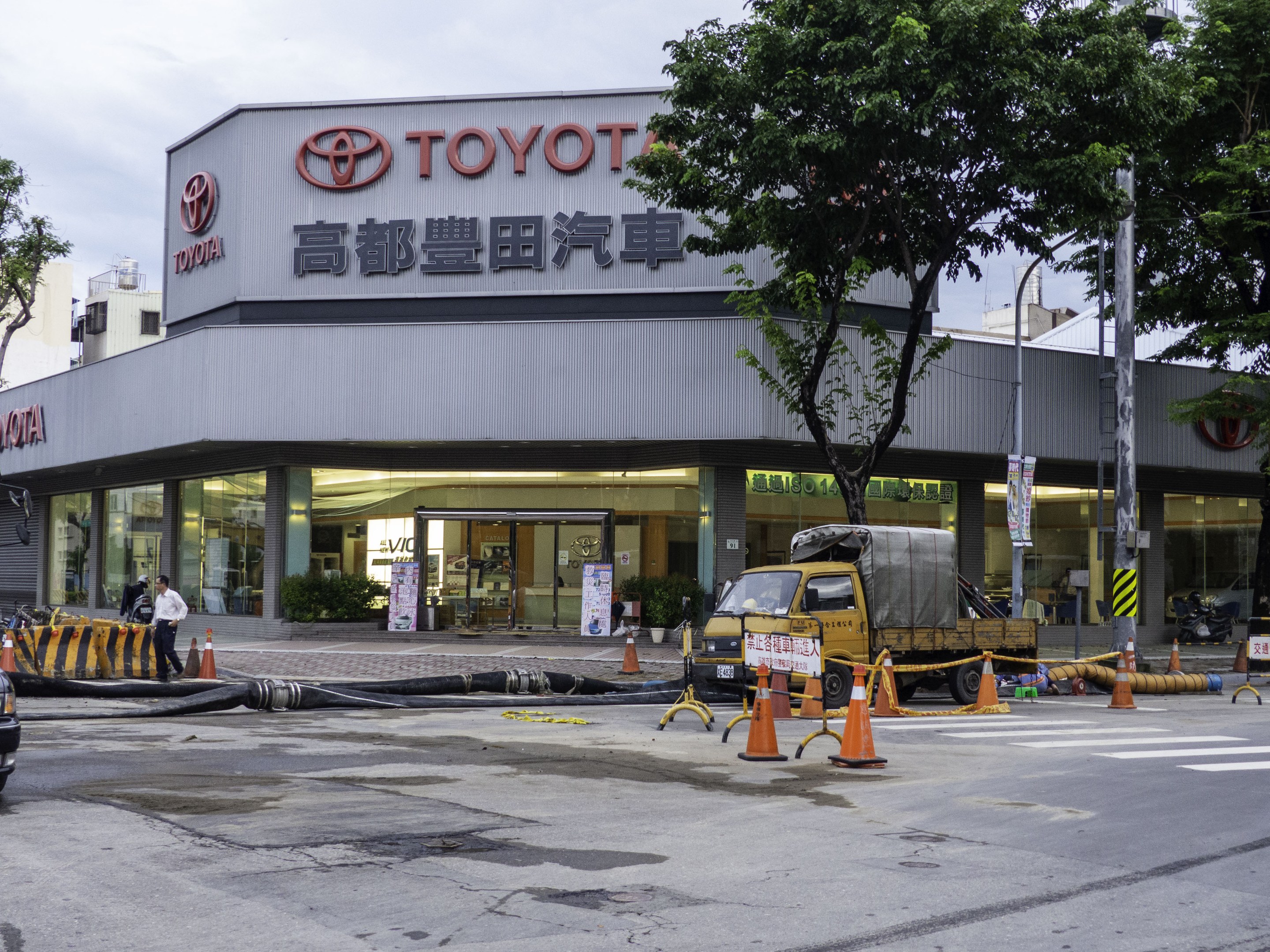 Ein Toyota-Autohaus in Tokyo, Japan, mit Fahrzeugen, Verkehrskegeln, Rohren, Menschen, Bäumen, Masten, Planken und Gebäuden im Vordergrund und einem bewülkten Himmel im Hintergrund.