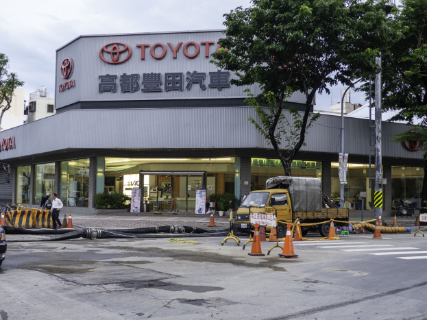 Ein Toyota-Autohaus in Tokyo, Japan, mit Fahrzeugen, Verkehrskegeln, Rohren, Menschen, Bäumen, Masten, Planken und Gebäuden im Vordergrund und einem bewülkten Himmel im Hintergrund.