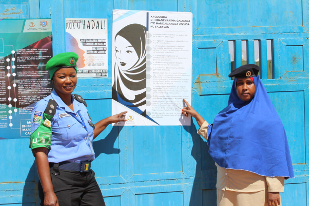 Zwei Frauen stehen nebeneinander vor einer blauen Tür mit Plakaten, eine trägt ein blaues Hemd, schwarze Hose und eine grüne Mütze, die andere hält ein Papier in der Hand.