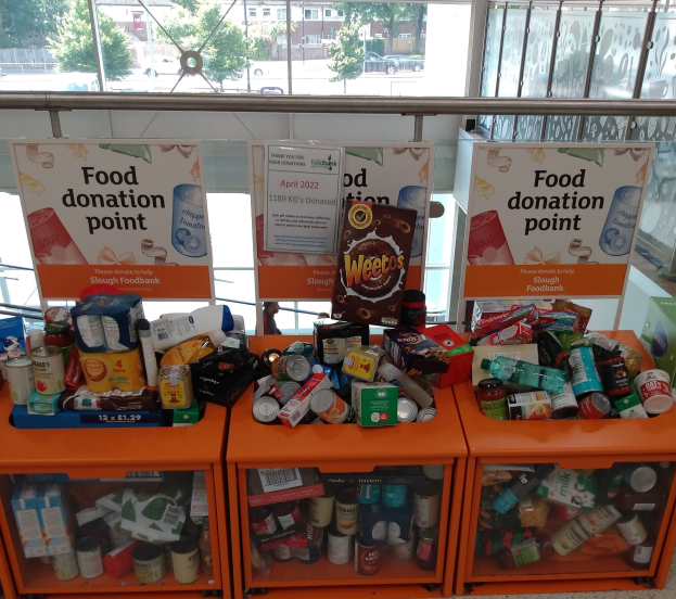 Orangene Schränke gefüllt mit Lebensmittelspenden wie Flaschen, Dosen und Kartons, mit Informationsschildern, durch Glasfenster betrachtet, die Outdoor-Fahrzeuge, Bäume, Gebäude und Himmel zeigen.