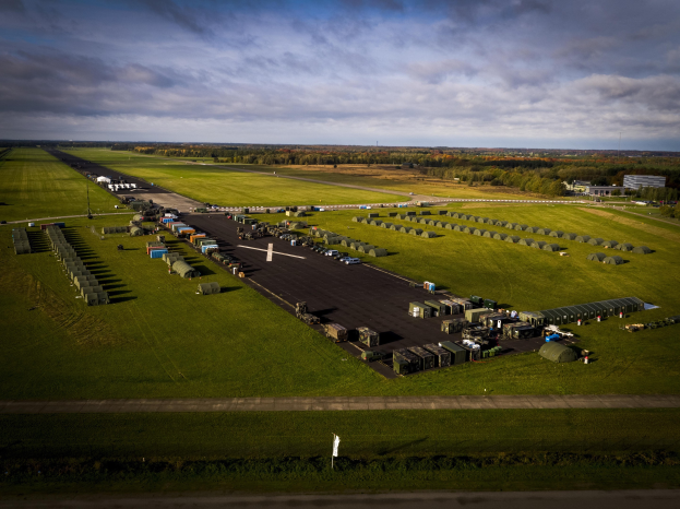 Luftaufnahme einer Militärbasis in den Niederlanden mit Fahrzeugen, Gebäuden, Bäumen, Gras und einem bewölkten Himmel.