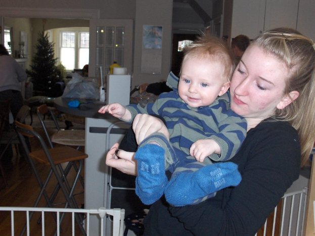 Eine Frau mit einem Baby auf der rechten Seite neben einem Gel├Ąnder, mit St├╝hlen im Hintergrund. Links steht eine Person bei einem Tisch mit vielen Gegenst├Ąnden. Im Hintergrund gibt es eine T├╝r, Fenster, einen Weihnachtsbaum und eine Wand mit etwas ├Ąnderem.