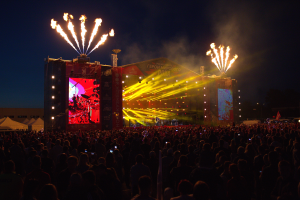 Eine große Menschenmenge auf einem Musikfestival mit einer hell erleuchteten Bühne, auf der Musiker spielen, Fahnen und Zelte links und Feuerwerk am dunklen Himmel.