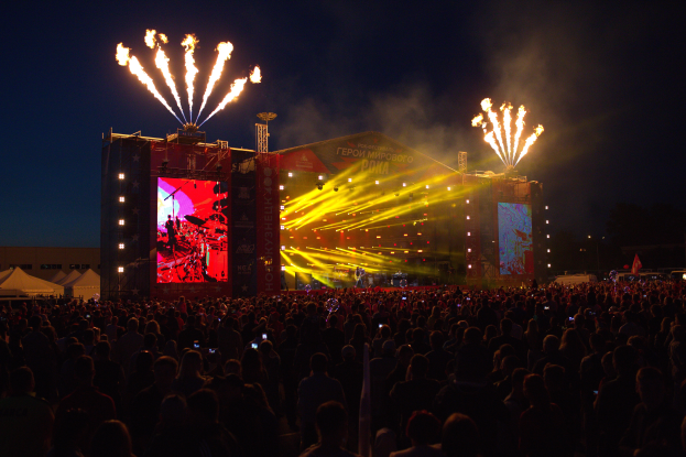Eine große Menschenmenge auf einem Musikfestival mit einer hell erleuchteten Bühne, auf der Musiker spielen, Fahnen und Zelte links und Feuerwerk am dunklen Himmel.