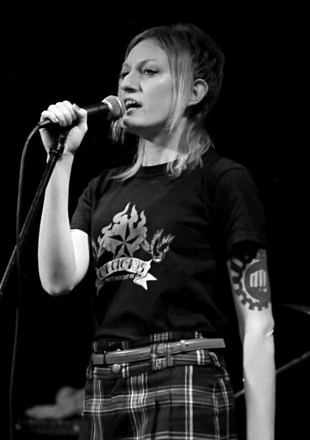 Eine Frau in schwarzer Bluse und kariertem Rock singt in ein Mikrofon mit einem Gürtel um die Taille, vor einem dunklen Hintergrund.