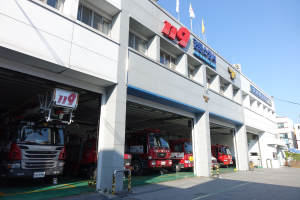 Eine Feuerwache mit mehreren Feuerwehrautos davor, mit Fenstern, Säulen und Namensschildern, sowie Fahnen im Vordergrund und anderen Gebäuden unter einem klaren blauen Himmel im Hintergrund.