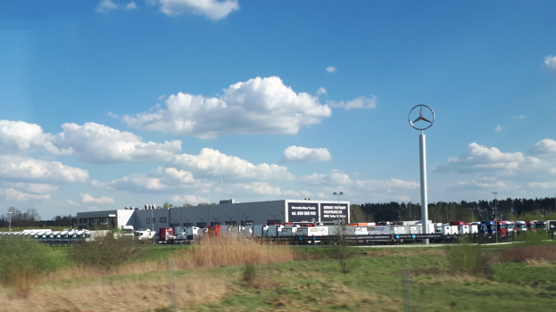 Ein Mercedes-Benz-Werk in Deutschland mit Fahrzeugen auf der Straße, umgeben von Gras, Pflanzen, Bäumen, Gebäuden und Polen, von denen einer ein Logo hat.