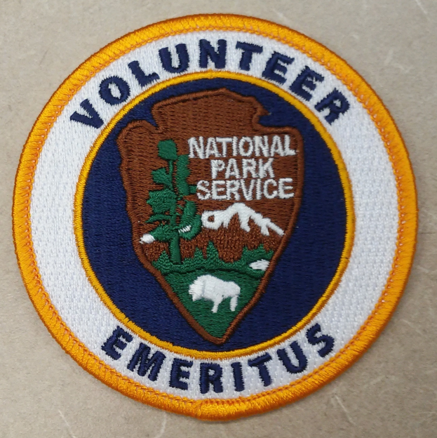 Ein kreisförmiges Flicken mit einem blauen Rand, das die Wörter 'Volunteer National Park Service Emeritus' in fetter schwarzer Schrift in der Mitte auf einem weißen Hintergrund zeigt, umgeben von einem gelben Kreis.