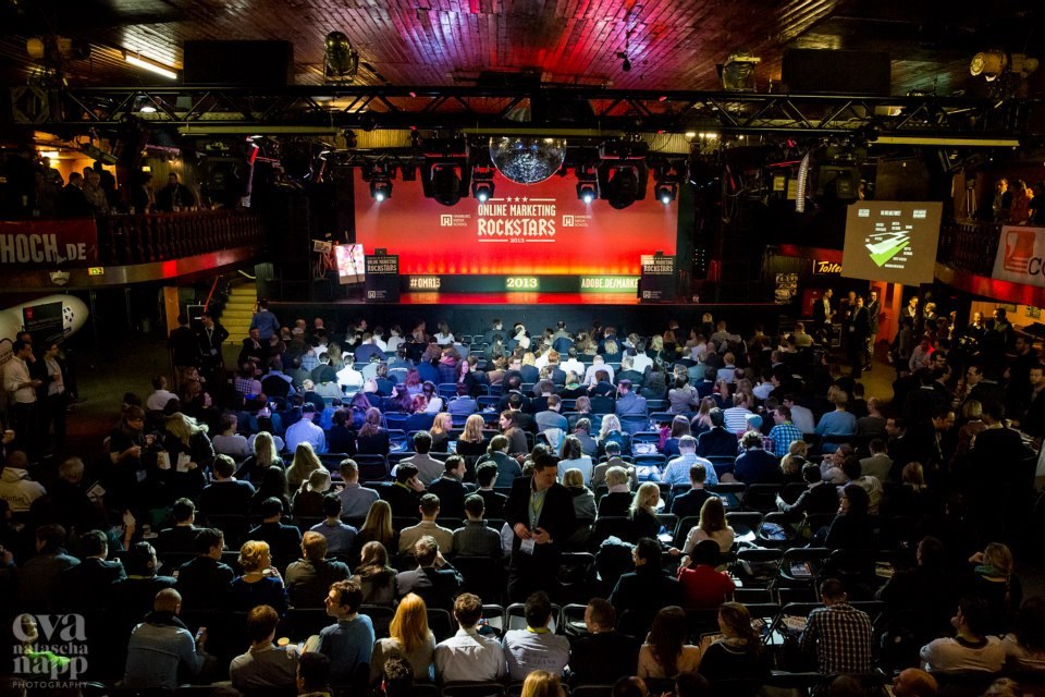 Eine große Menschenmenge sitzt auf Stühlen, die einer hell erleuchteten Bühne mit Bildschirmen und Bannern gegenüberstehen und auf ein Ereignis warten.