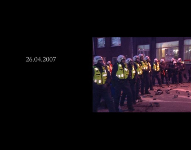 Eine Gruppe von Polizisten in Helmen und Jacken steht vor einem Gebäude mit Fenstern, wobei auf der linken Seite des Bildes ein Datum zu sehen ist.