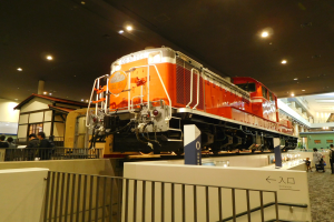 Ein roter und weißer Zug in einem Museum ausgestellt, umgeben von Menschen, einem Zaun, einem Schild, Pfosten, einem Dach mit Deckenleuchten und Gebäuden.