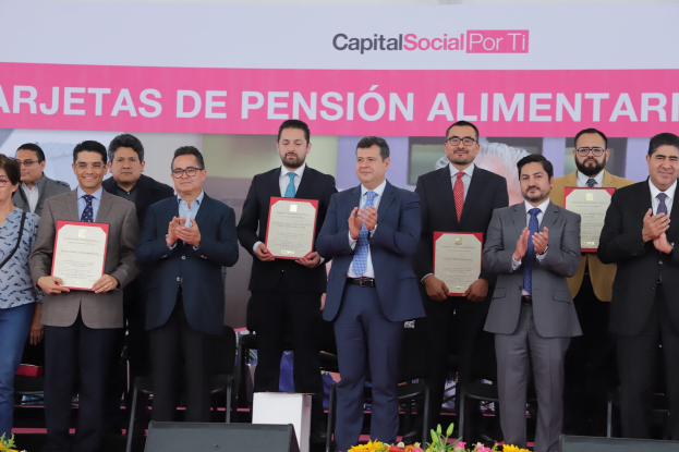 Eine Gruppe von Menschen auf einer Bühne, die klatschen und Zertifikate halten, mit Stühlen in einem Halbkreis angeordnet, ein Banner mit der Aufschrift "Capital Social for Ti" im Hintergrund und Blumensträuße und Lautsprecher unten.